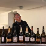 oito garrafas diferentes de espumante em linha, com a sommelier Flávia de Carvalho por trás, a mostrar os rótulos que foram provados neste dia do encontro nacional de espumantes, Portugal